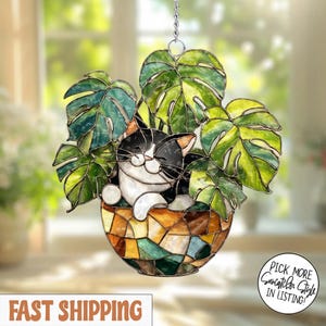 May include: Stained glass suncatcher featuring a black and white cat nestled in a pot of green monstera leaves. The pot has a mosaic design with shades of brown, green, and blue. A silver chain is attached for hanging.