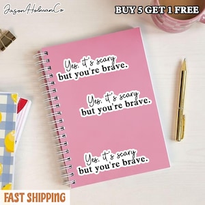 Puede incluir: Un cuaderno de espiral rosa con tres pegatinas blancas id&eacute;nticas con el texto en negro, Yes. it's scary but you're brave. El cuaderno est&aacute; colocado junto a un bol&iacute;grafo dorado y una parte de un cuaderno de cuadros azules y blancos.