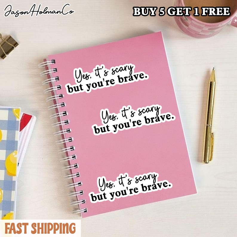 Puede incluir: Un cuaderno de espiral rosa con tres pegatinas blancas id&eacute;nticas con el texto en negro, Yes. it's scary but you're brave. El cuaderno est&aacute; colocado junto a un bol&iacute;grafo dorado y una parte de un cuaderno de cuadros azules y blancos.