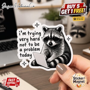 May include: A die-cut sticker featuring a black and white illustration of a raccoon wearing glasses and typing on a laptop. The text reads, i'm trying very hard not to be a problem today, surrounded by small four-pointed star icons on a white background.