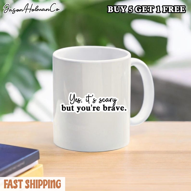 Puede incluir: Una taza de cer&aacute;mica blanca con el texto impreso en negro Yes. it's scary but you're brave. La taza tiene una forma cil&iacute;ndrica est&aacute;ndar con un asa curva en el lado derecho, presentada sobre un fondo borroso con una pila de libros.