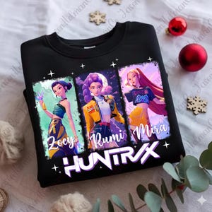 May include: Black sweatshirt featuring three stylized female figures with the names Zoey, Rumi, and Mira. The sweatshirt has the word "HUNTRAX" in white block letters. The figures are in colorful, artistic renderings.