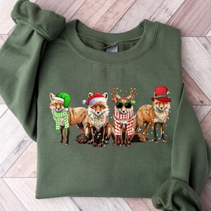 Può includere: Felpa verde con un design natalizio con quattro volpi che indossano cappelli e sciarpe festivi. Le volpi sono circondate da luci natalizie.