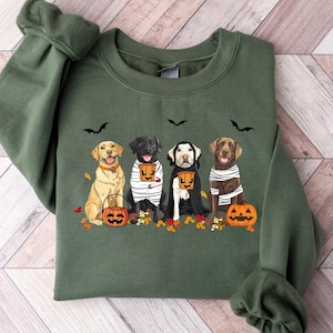 May include: A green sweatshirt with a Halloween-themed design featuring four Labrador Retrievers dressed in costumes and holding pumpkins. The dogs are wearing black and white striped shirts and have orange pumpkins in their paws.