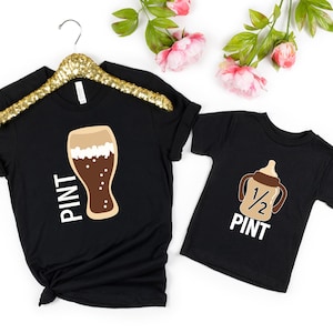 以下が含まれることがあります： ビールグラスと哺乳瓶のグラフィックが描かれた黒いTシャツ2枚。大人のTシャツには「PINT」、ベビーTシャツには「1/2 PINT」と書いてあります。