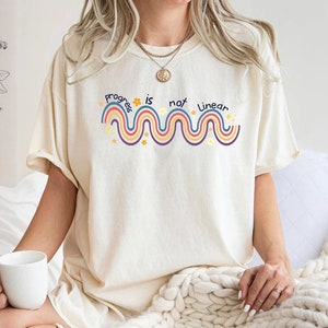 Puede incluir: Una camiseta blanca con un diseño gráfico que dice "Progress is not linear" con un diseño tipo arcoíris y estrellas. La camiseta la lleva puesta una persona sentada en una cama blanca.