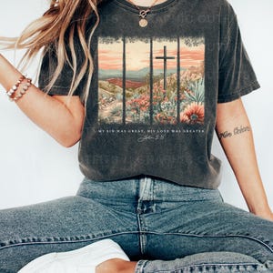 Puede incluir: Una camiseta gris oscuro con un gr&aacute;fico de estilo vintage de una cruz en un paisaje des&eacute;rtico. El texto "My sin was great, His love was greater. John 3:16" est&aacute; impreso debajo del gr&aacute;fico.