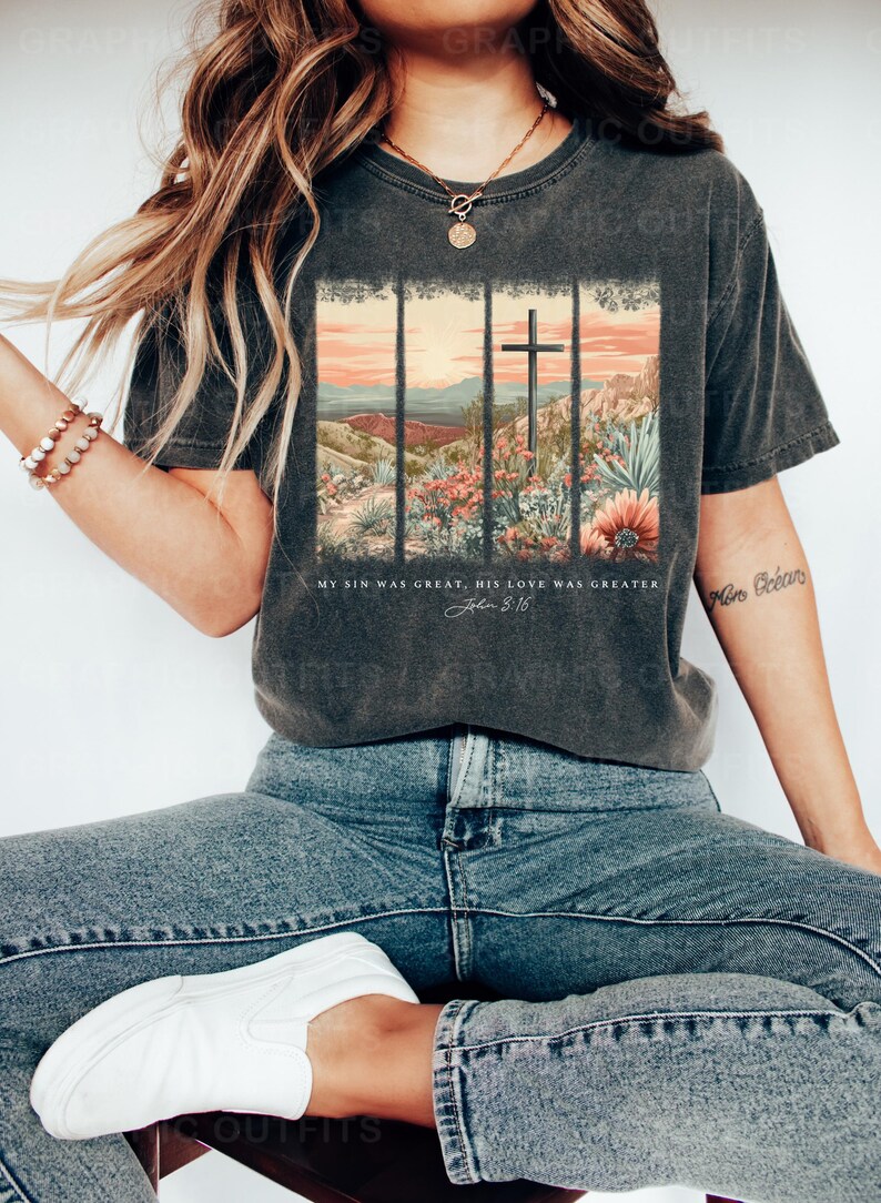 Puede incluir: Una camiseta gris oscuro con un gr&aacute;fico de estilo vintage de una cruz en un paisaje des&eacute;rtico. El texto "My sin was great, His love was greater. John 3:16" est&aacute; impreso debajo del gr&aacute;fico.