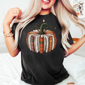 May include: Black t-shirt featuring a pumpkin design made of open books. The pumpkin has a green stem and is composed of various book spines in shades of orange, red, and brown. The open pages of the books are visible, creating a unique fall-themed graphic.