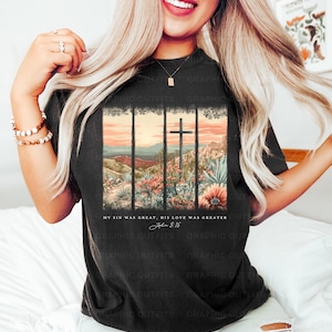 Puede incluir: Una camiseta negra con un gr&aacute;fico de un paisaje de puesta de sol con una cruz en el centro. El texto "My sin was great, His love was greater John 8:16" est&aacute; impreso debajo del gr&aacute;fico.