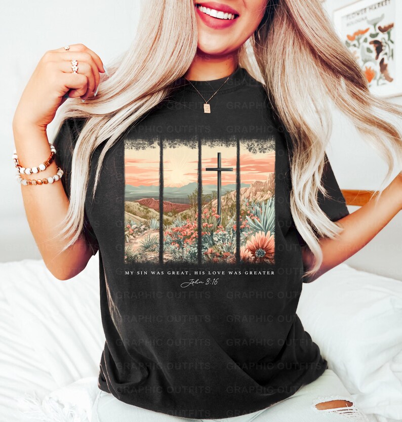 Puede incluir: Una camiseta negra con un gr&aacute;fico de un paisaje de puesta de sol con una cruz en el centro. El texto "My sin was great, His love was greater John 8:16" est&aacute; impreso debajo del gr&aacute;fico.