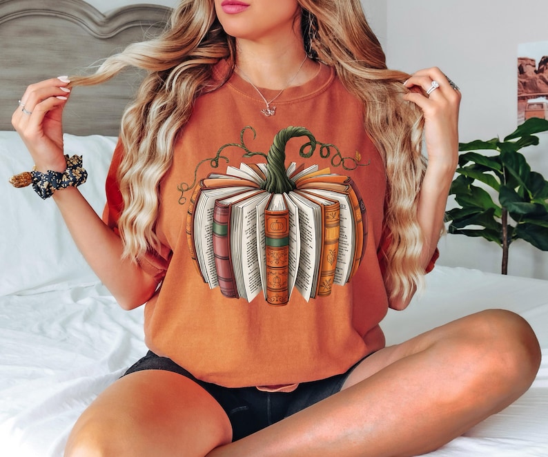 May include: Orange t-shirt featuring a pumpkin design made of open books with a green stem and vines. The graphic tee is a comfortable, casual style, perfect for fall and book lovers.