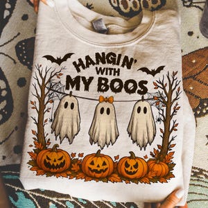 Op de afbeelding: Een lichtbeige sweatshirt met een Halloween-ontwerp. Het ontwerp bevat drie cartoongeesten, pompoenen, vleermuizen en de tekst "Hangin' with my Boos".