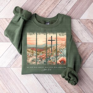 Puede incluir: Una sudadera verde con un dise&ntilde;o de cruz y flores. El texto en la sudadera dice "My sin was great, His love was greater John 3:16".
