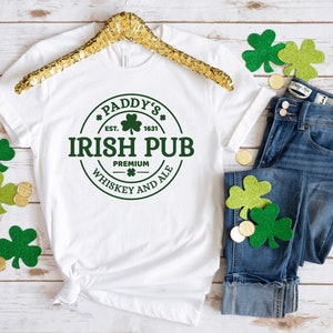 May include: White t-shirt with a green circle graphic that says "Paddy's Irish Pub Est. 1631 Premium Whiskey and Ale". The shirt is on a wooden surface with a gold hanger and green shamrock decorations.
