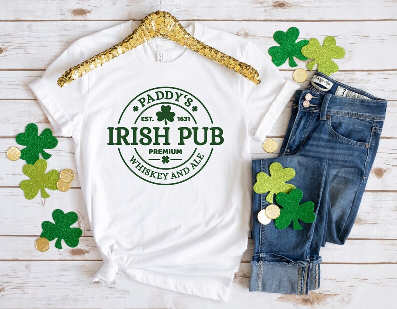May include: White t-shirt with a green circle graphic that says "Paddy's Irish Pub Est. 1631 Premium Whiskey and Ale". The shirt is on a wooden surface with a gold hanger and green shamrock decorations.