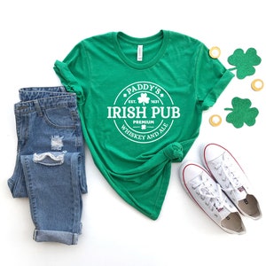 May include: Green t-shirt with a white circle graphic that says "Paddy's Irish Pub Est. 1831 Premium Whiskey and Ale". The shirt is paired with blue jeans and white sneakers.