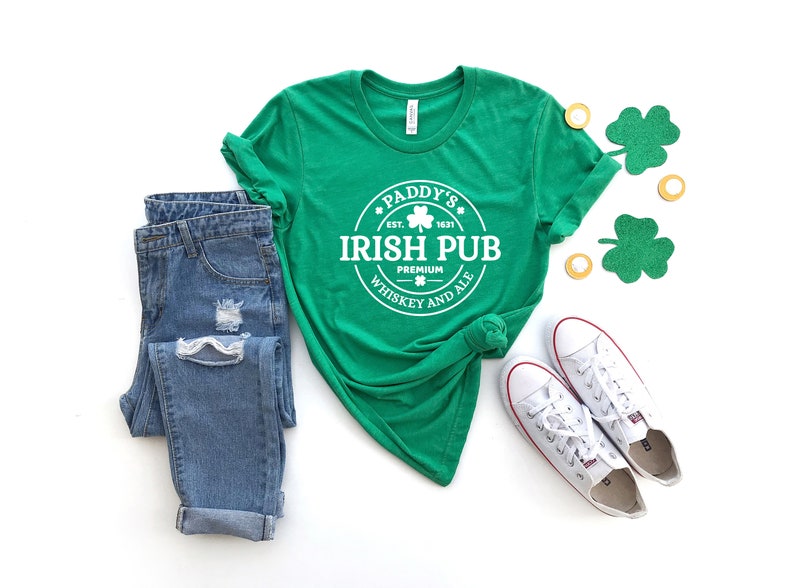May include: Green t-shirt with a white circle graphic that says "Paddy's Irish Pub Est. 1831 Premium Whiskey and Ale". The shirt is paired with blue jeans and white sneakers.