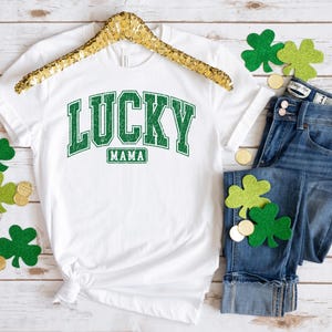 May include: White t-shirt with green text that says "LUCKY MAMA" in a vintage collegiate style font. The shirt is on a wooden surface with green shamrocks and gold coins.