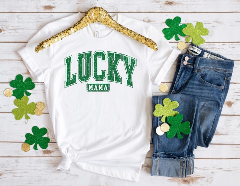 May include: White t-shirt with green text that says "LUCKY MAMA" in a vintage collegiate style font. The shirt is on a wooden surface with green shamrocks and gold coins.