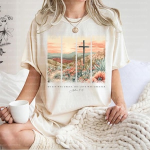 Puede incluir: Una camiseta beige con un gr&aacute;fico de una cruz en un paisaje des&eacute;rtico. El texto "My sin was great, His love was greater, John 3:16" est&aacute; impreso debajo del gr&aacute;fico.