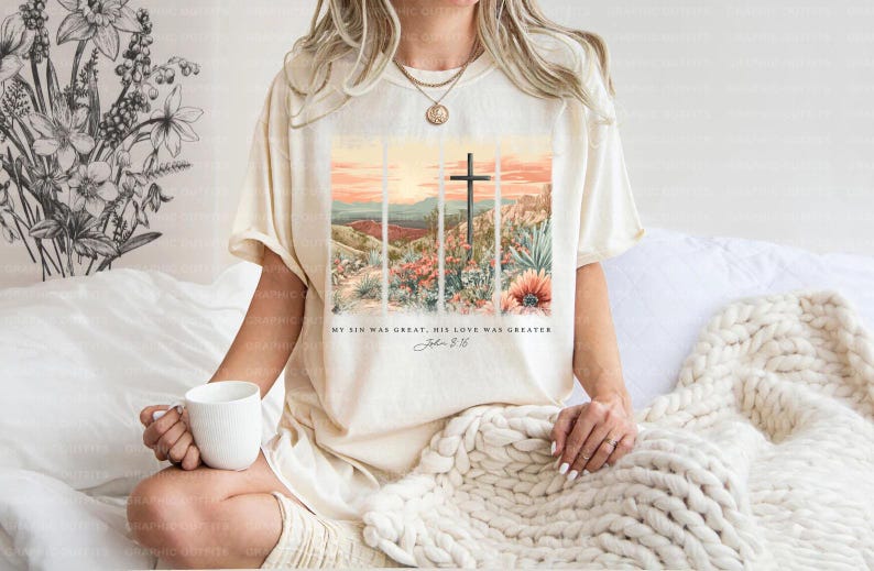 Puede incluir: Una camiseta beige con un gr&aacute;fico de una cruz en un paisaje des&eacute;rtico. El texto "My sin was great, His love was greater, John 3:16" est&aacute; impreso debajo del gr&aacute;fico.