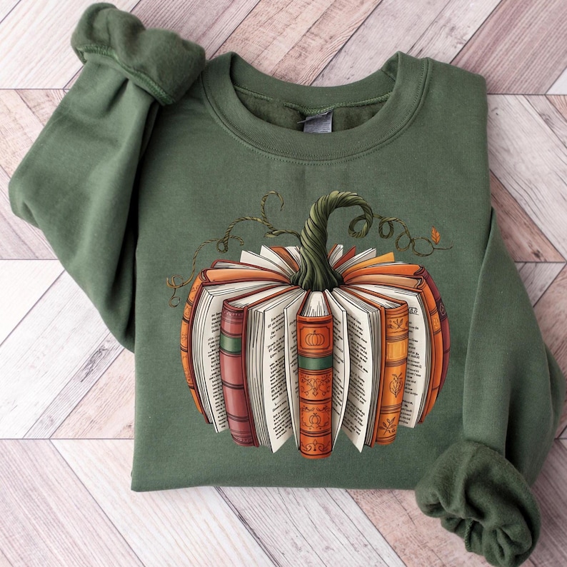 May include: Green sweatshirt featuring a pumpkin design made of open books. The pumpkin has a green stem and vine details. The books are orange, red, and brown with white pages. A fall-themed graphic for book lovers.
