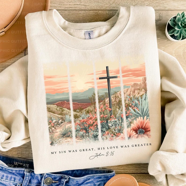 Puede incluir: Una sudadera beige con un estampado gr&aacute;fico de una cruz en un paisaje des&eacute;rtico con el texto "My sin was great, His love was greater John 3:16".