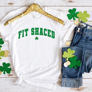 May include: White t-shirt with the text "Fit Shaced" in green letters with a shamrock underneath. The shirt is on a hanger and there are shamrocks and gold coins scattered around it.