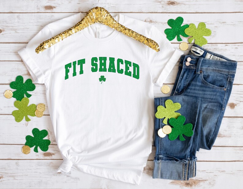 May include: White t-shirt with the text "Fit Shaced" in green letters with a shamrock underneath. The shirt is on a hanger and there are shamrocks and gold coins scattered around it.