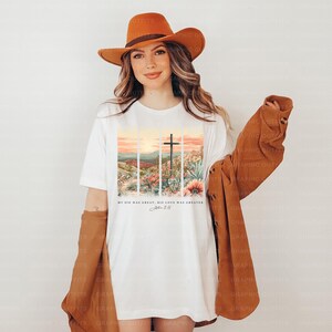 Puede incluir: Camiseta blanca con un gr&aacute;fico de una cruz y un paisaje de puesta de sol. El texto "My sin was great, His love was greater" y "John 3:16" est&aacute; impreso en la camiseta.