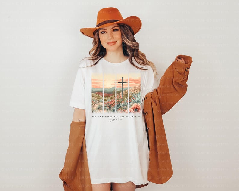 Puede incluir: Camiseta blanca con un gr&aacute;fico de una cruz y un paisaje de puesta de sol. El texto "My sin was great, His love was greater" y "John 3:16" est&aacute; impreso en la camiseta.