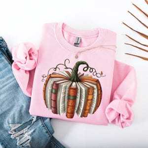 May include: A light pink sweatshirt featuring a pumpkin design made of books. The pumpkin has a green stem and is surrounded by orange, brown, and white books. A silver necklace is draped across the sweatshirt, which is paired with ripped blue jeans.