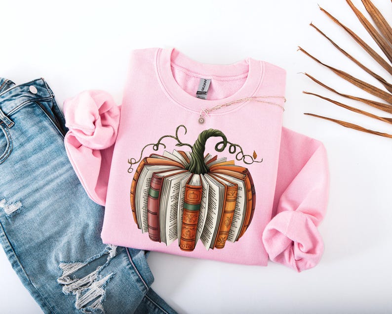 May include: A light pink sweatshirt featuring a pumpkin design made of books. The pumpkin has a green stem and is surrounded by orange, brown, and white books. A silver necklace is draped across the sweatshirt, which is paired with ripped blue jeans.