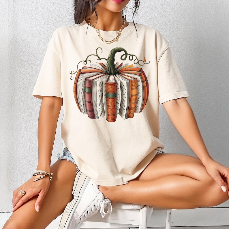 May include: Cream-colored t-shirt featuring a pumpkin design made of books. The pumpkin has a green stem and is composed of open books with colorful spines. The design is centered on the shirt, creating a fall-themed aesthetic.