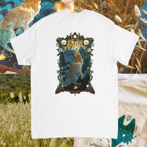 May include: A white t-shirt featuring a graphic print of a wolf howling at a glowing sun within an ornate, dark-framed border decorated with vines and flowers. A smaller print on the fabric includes a stylised wolf head and the text PROTECT YOUR LIGHT.