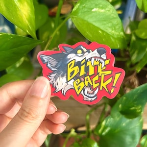 May include: A die-cut sticker featuring a stylised illustration of a snarling wolf head with yellow eyes and bared teeth. The text BITE BACK! is printed in bold yellow letters across the centre of the wolf, all set against a vibrant red background.