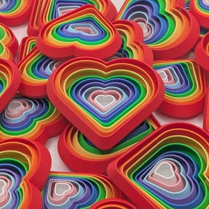 May include: A selection of multi-coloured heart-shaped cookie cutters. Each cutter has a red outer edge and a rainbow of inner layers, creating a nested effect. The cutters are arranged in a scattered pattern, displaying their vibrant colours. Metric measurements.