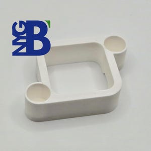 Op de afbeelding: Een wit, vierkant object met twee cilindrische uitsteeksels. Het object heeft een hol midden en afgeronde randen. De letters "NMG" en "B" zijn linksboven in blauw en groen gedrukt. Het object staat op een witte achtergrond.