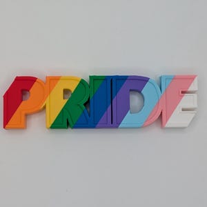 Op de afbeelding: Een driedimensionaal bord dat het woord PRIDE spelt in blokletters. De letters hebben een diagonaal gestreept patroon met kleuren zoals rood, oranje, geel, groen, blauw, paars, lichtblauw, roze en wit over het gehele oppervlak van het object.