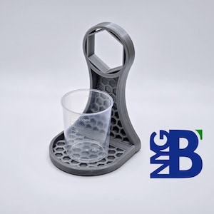 Puede incluir: Un soporte gris impreso en 3D con una abertura hexagonal en la parte superior y una taza pequeña en una plataforma. El soporte está diseñado para sostener una taza pequeña y una llave.