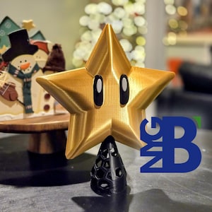 May include: A gold star-shaped ornament with black eyes and a black base. The star is a 3D-printed item. A decorative snowman and Christmas tree are in the background. The star is approximately 15 cm tall.
