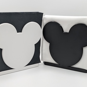 Può includere: Due portatovaglioli, uno con un design a testa di Topolino bianco e tovaglioli neri, l'altro con un design a testa di Topolino nero e tovaglioli bianchi. I supporti sono rettangolari con una base.
