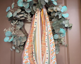May include: A circular wreath constructed from brown twigs and adorned with teal-tinted eucalyptus leaves and sprigs. A collection of long, hanging ribbons in various patterns, including orange and teal pumpkins, autumn leaves, and stripes, hangs down.