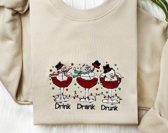 Pode incluir: Camisola bege de gola redonda com um desenho bordado de três bonecos de neve em taças de vinho. Os bonecos de neve usam chapéus e cachecóis, com as palavras "Drink Drank Drunk" por baixo. Roupa festiva de férias.