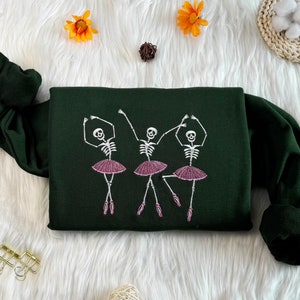 May include: A dark green sweatshirt featuring a white embroidered design of three skeletons dressed as ballerinas. The skeletons are wearing pink tutus and are in various dance poses.