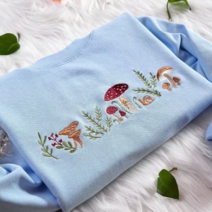 Op de afbeelding: Een lichtblauw sweatshirt met een geborduurd ontwerp van paddenstoelen, bladeren en een slak. Het ontwerp is in tinten bruin, rood en groen.