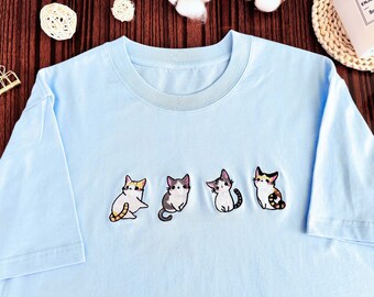 May include: A light blue t-shirt with four embroidered cats in different poses. The cats are white, gray, and brown with black eyes.
