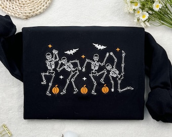 May include: Black sweatshirt featuring an embroidered design of dancing skeletons, pumpkins, and bats. The design is in white and orange. The sweatshirt has long sleeves and is folded. The background includes white flowers and cotton buds.