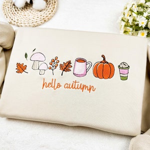 Op de afbeelding: Een beige sweatshirt met een geborduurd ontwerp met een paddenstoel, bladeren, een kopje koffie, een pompoen en een koffiebeker. De tekst "hello autumn" is onder het ontwerp geborduurd.